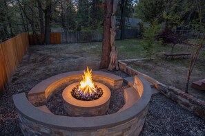 A circular stone fire pit with a fire burning in the center, surrounded by a wooden fence and trees. - Powder & Pines. Hot Tub, Sauna & Walk to Village (Big Bear Lake)