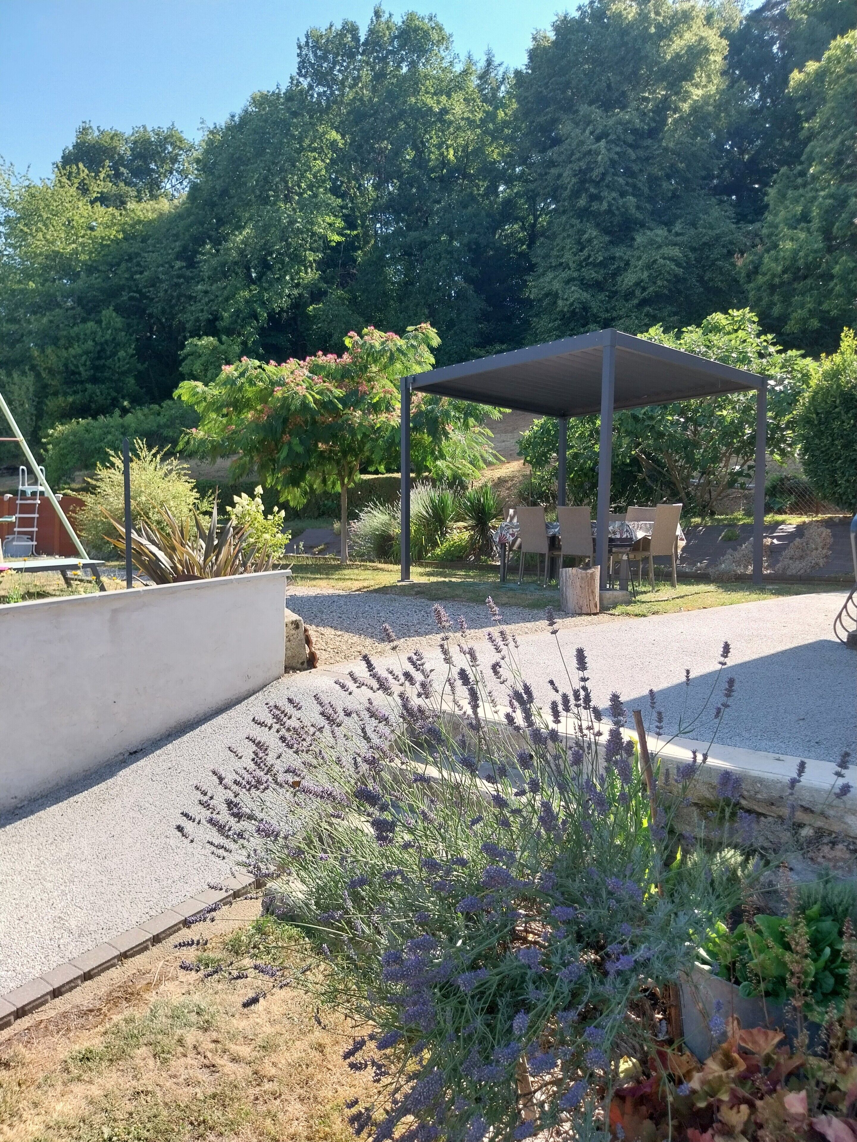 A covered outdoor seating area with a gravel path, surrounded by various plants and trees.