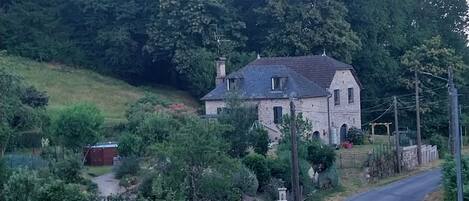 A stone house with a sloped roof, surrounded by a fenced yard and a curved road.