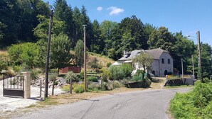A rural road leading to a stone house with a sloped roof, surrounded by dense greenery and trees.