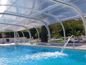 Ein überdachter Pool mit Glasdach, Liegestühlen und einem Wasserspiel.