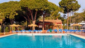 Ein Swimmingpool mit blauen Liegestühlen und Sonnenschirmen, umgeben von Bäumen und einem Gebäude mit rotem Dach.