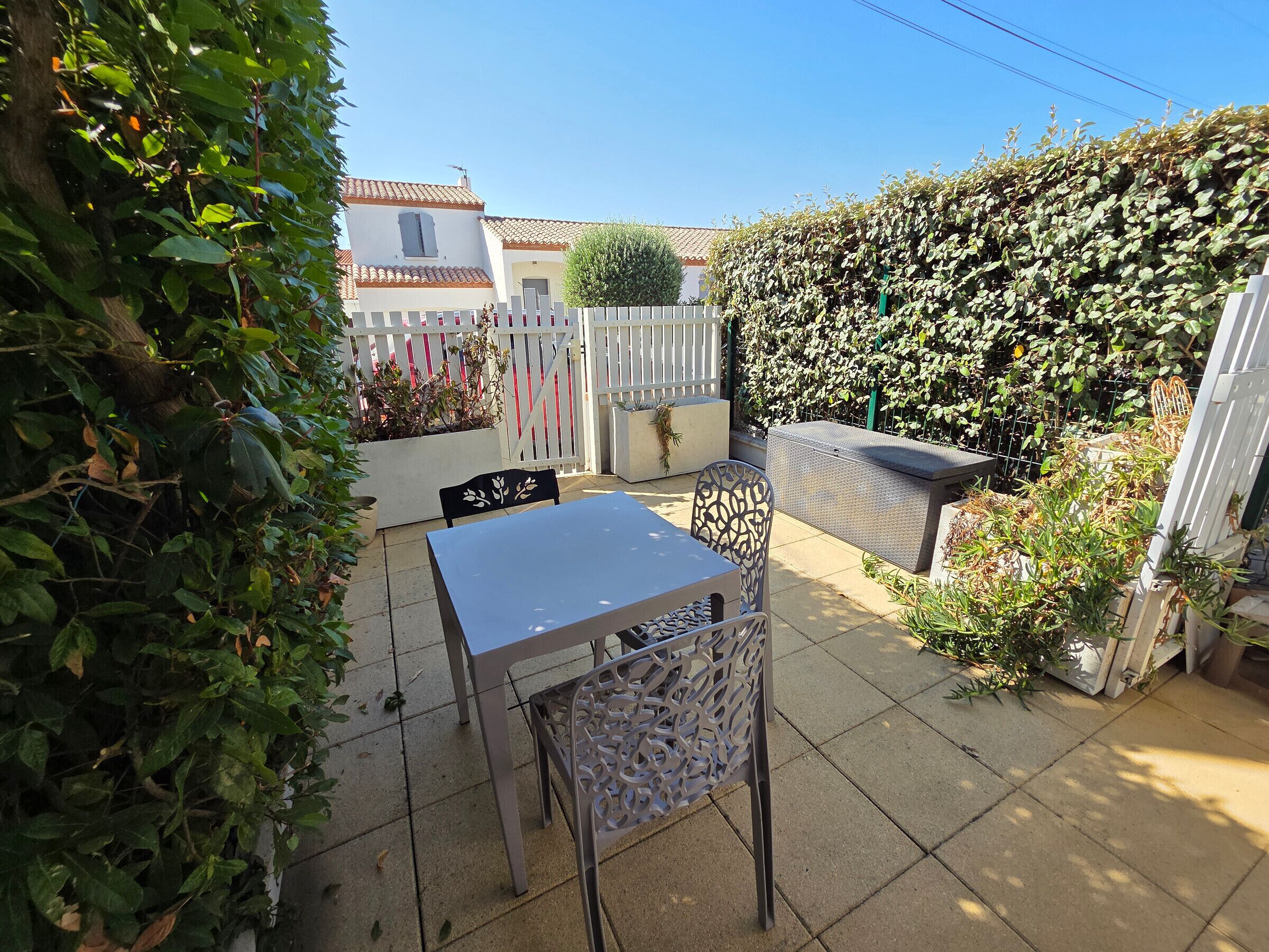 A patio with a table and chairs, surrounded by a white fence and dense foliage.