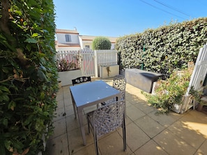 A patio with a table and chairs, surrounded by a white fence and dense foliage.