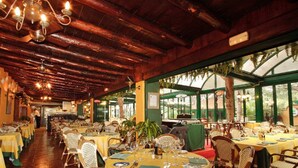 Een restaurant met houten balken en kroonluchters, tafels gedekt met gele tafelkleden en rieten stoelen, en uitzicht op groen buiten.