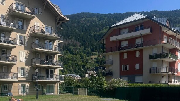 Una piscina amb un pont de fusta, envoltada d'edificis d'altes plantes amb balcons, amb un paisatge de muntanyes al fons.