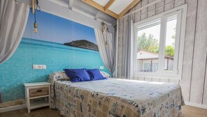 A bedroom with a bed, a nightstand, a mural of a beach scene, and a window with a view of greenery. - Bungalow with Terrace and Accessible Ramp in Guardamar (Guardamar del Segura)