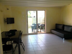 Ein Wohnzimmer mit einer Couch, einem Esstisch mit Stühlen, einem an der Wand befestigten Fernseher und einer Glastür zum Außenbereich mit Sitzgelegenheiten.