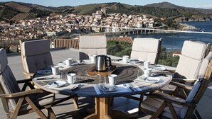 Ein runder Tisch mit Stühlen, einer Kaffeekanne und Besteck auf einer Terrasse mit Blick auf eine Küstenstadt.
