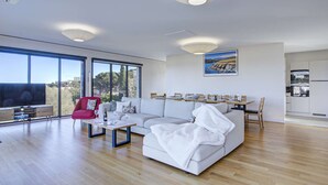 Ein modernes Wohnzimmer mit einem großen Sofa, einem roten Sessel, einem Couchtisch und einem Fernseher. Große Fenster bieten einen Blick ins Freie.