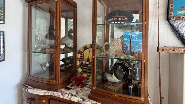 Une vitrine en bois aux portes vitrées, contenant divers objets, notamment une roue de navire, une maquette de bateau et une enseigne portant l'inscription « Marine Nationale ».