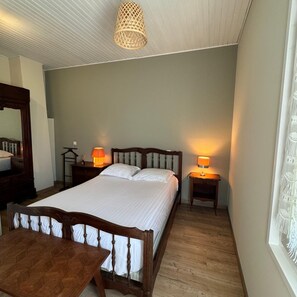 Un dormitorio con una cama de madera, dos mesitas de noche con lámparas, una silla de madera, una mesa de madera y un candelabro colgante.
