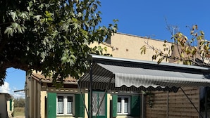 Un petit espace extérieur aménagé pour les repas, avec une table et des chaises, une terrasse couverte et un bâtiment aux volets verts.