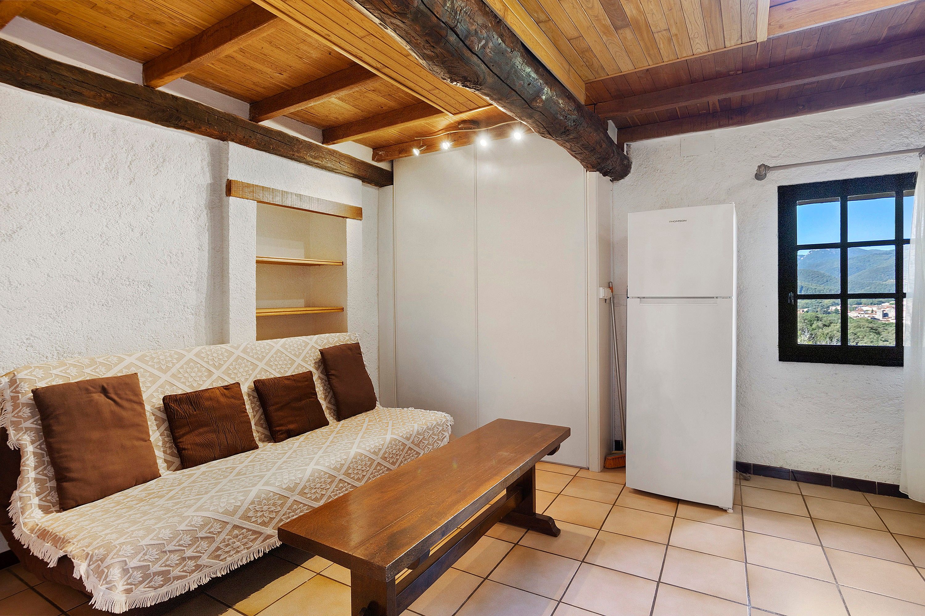 Une pièce avec un banc en bois, un canapé blanc avec des coussins marron, un réfrigérateur blanc et une fenêtre donnant sur un paysage agréable.