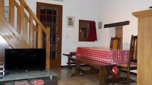 A living room with a TV, books, a wooden staircase, a window with curtains, and a dining table with chairs.