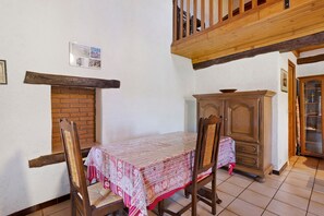 A dining area with a table covered in a patterned tablecloth, two wooden chairs, a brick fireplace, a wooden cabinet, and a staircase leading to an upper level. - Holiday Home 'Le Mas' with Mountain View, Wi-Fi, Air Conditioning (Arles-sur-Tech)