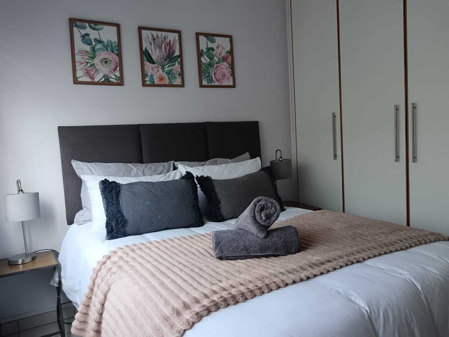 A neatly made bed with a grey headboard, white and grey pillows, and a beige blanket. There are three framed floral paintings on the wall above the bed.