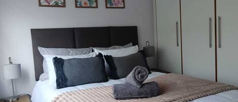 A neatly made bed with a grey headboard, white and grey pillows, and a beige blanket. There are three framed floral paintings on the wall above the bed.