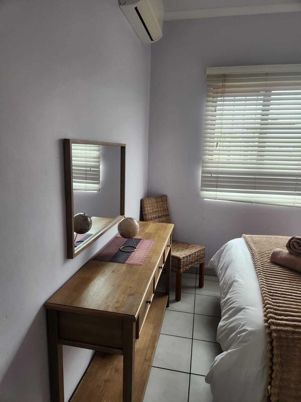 A bedroom with a wooden desk, a chair, a bed with a rolled towel, a window with blinds, and a wall-mounted air conditioner.