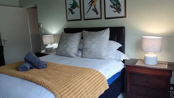 Una cama bien hecha con una manta color beige, una mesita de noche con lámpara, y tres cuadros con pájaros en la pared.