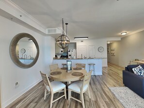 A modern kitchen with white cabinets, a central island, and a dining area with a round table and chairs. - Tidewater Beach Resort #503 | Gulf Front + Amenities (Panama City Beach)