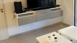 Uma sala de estar moderna com uma televisão de tela plana fixada em um armário branco, duas mesas brancas e uma porta à esquerda.
