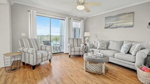 A spacious living room with a large window, a ceiling fan, and a grey sofa. - Cozy 2-bedroom condo at Myrtle Beach Resort (Myrtle Beach)