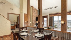 Uma sala de jantar com uma mesa de madeira posta para quatro pessoas, rodeada por cadeiras. Uma parede de destaque em tijolo e vista para o exterior são visíveis.