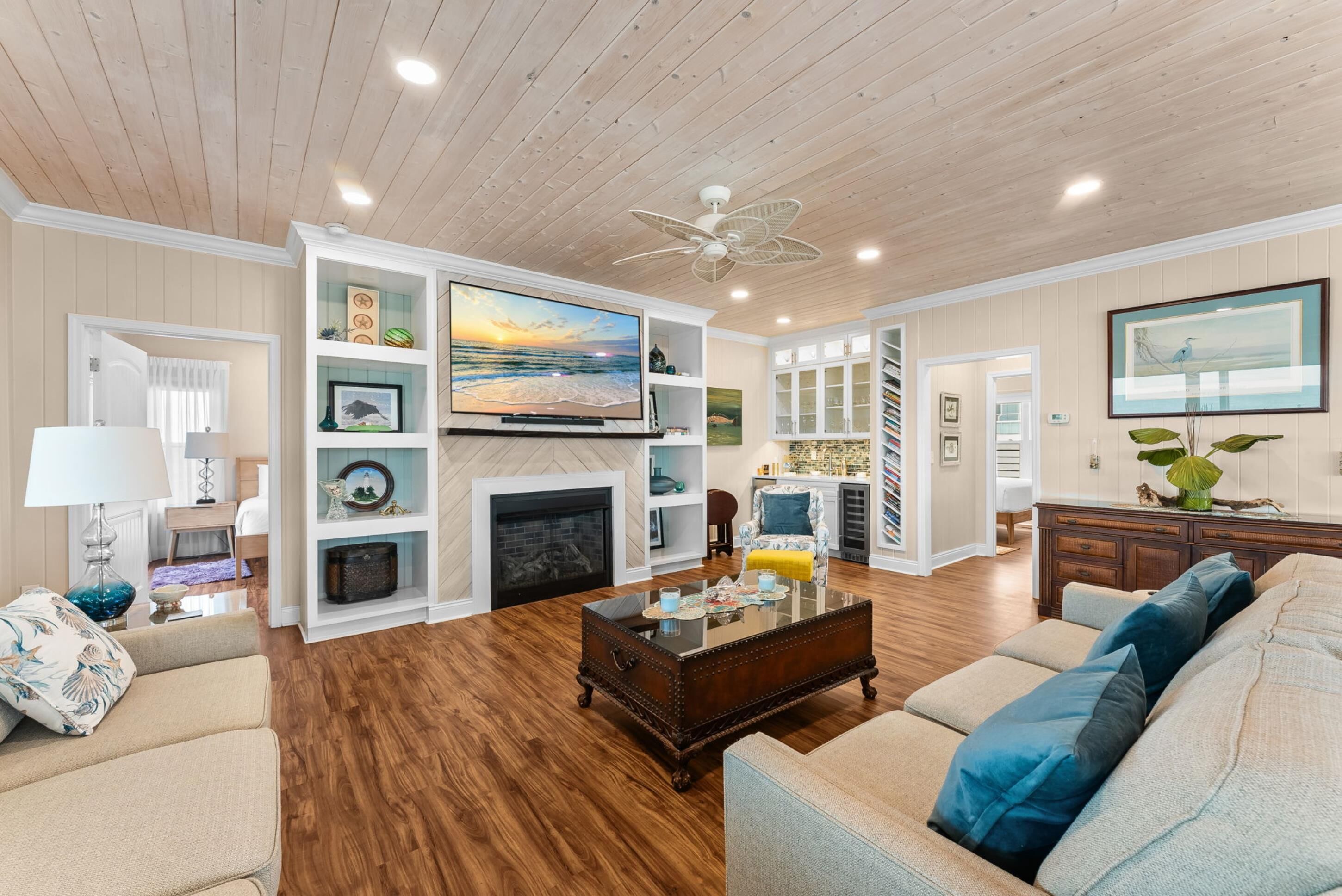 A living room with a fireplace, wooden flooring, a ceiling fan, and a large TV.