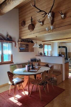 A rustic interior with a wooden dining table, chairs, and a red rug. There are antlers mounted on the wall and a kitchen area with a sink and cabinets. - Alpe Monte Massella by Interhome (Poschiavo)