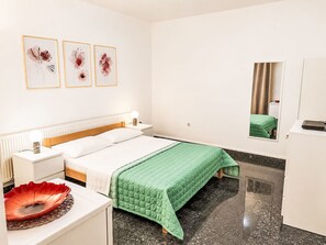 Et hotelværelse med en seng, et natbord, en lampe, grønt sengetæppe, kunst på væggen og et spejl.