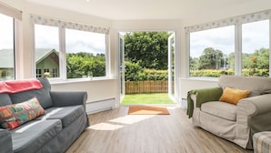 A spacious living room with two sofas, a wooden floor, and large windows offering a view of a garden and neighboring houses. - Ammerham Farm Cottage (Chard)