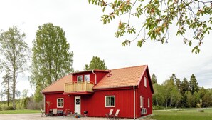 A red wooden house with a balcony, a gravel driveway, and a grassy yard.
