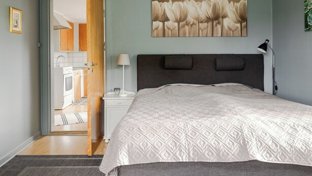 A bedroom with a bed, a nightstand, a lamp, a painting, and a view into the kitchen.