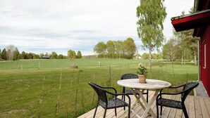 A wooden deck with a round table and four chairs, overlooking a grassy field with trees and a distant building.