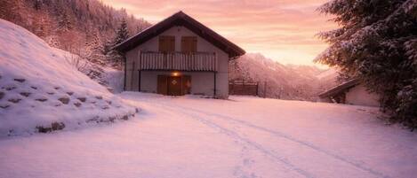A wooden cabin with a balcony, surrounded by snow-covered trees and a clear sky.