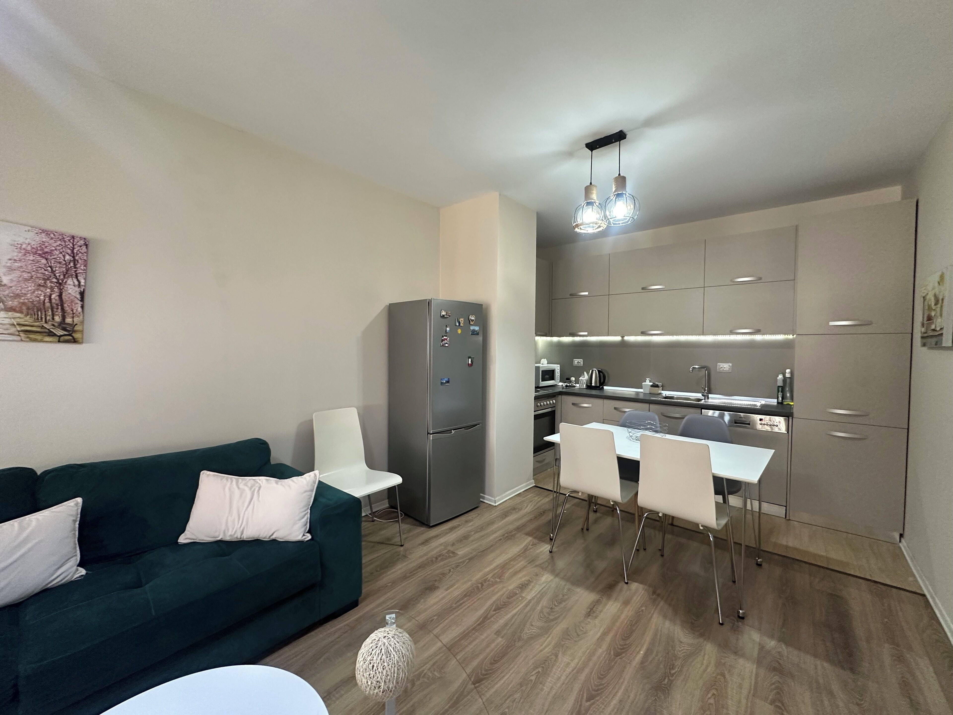 A modern living room with a green sofa, a dining table with chairs, and a kitchen area with stainless steel appliances and wooden flooring.
