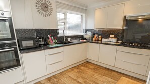 A modern kitchen with white cabinets, a black countertop, a microwave, a stove, and a window with blinds. - Red Brick Haven (East Cowes)
