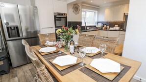 A modern kitchen with stainless steel appliances, a dining area with a long wooden table set for four, and a wicker chair. - Red Brick Haven (East Cowes)