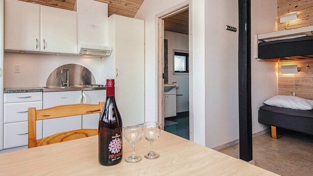 A compact kitchen with white cabinets, a stainless steel sink, and a wooden ceiling. A dining table with a wine bottle and glasses is in the foreground. A bunk bed is visible in the background. - Standard Cabin For, 5 Pers, 25m2 (Ronde)