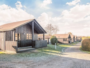 Een rij houten hutten met veranda en overdekte gangpad, gelegen in een landelijke omgeving met een grindpad en schaarse begroeiing.