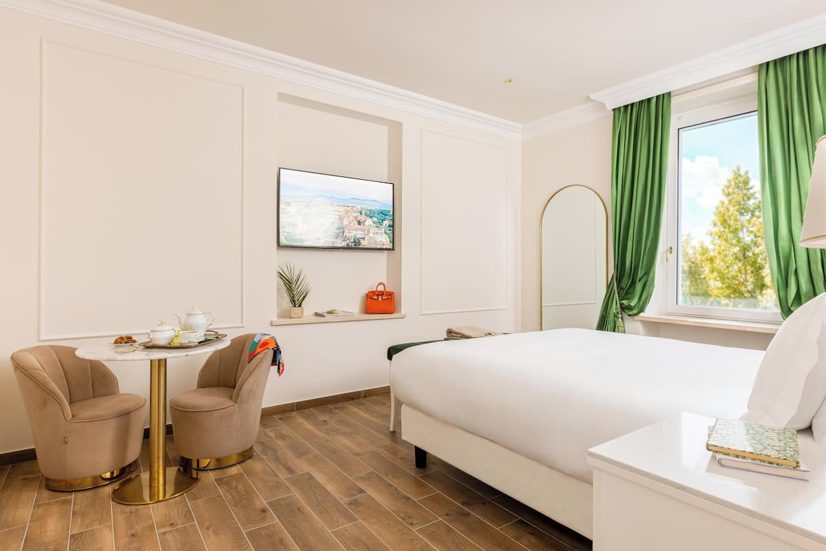 A hotel room with a bed, a small round table with a chair, a TV on the wall, and a window with curtains.