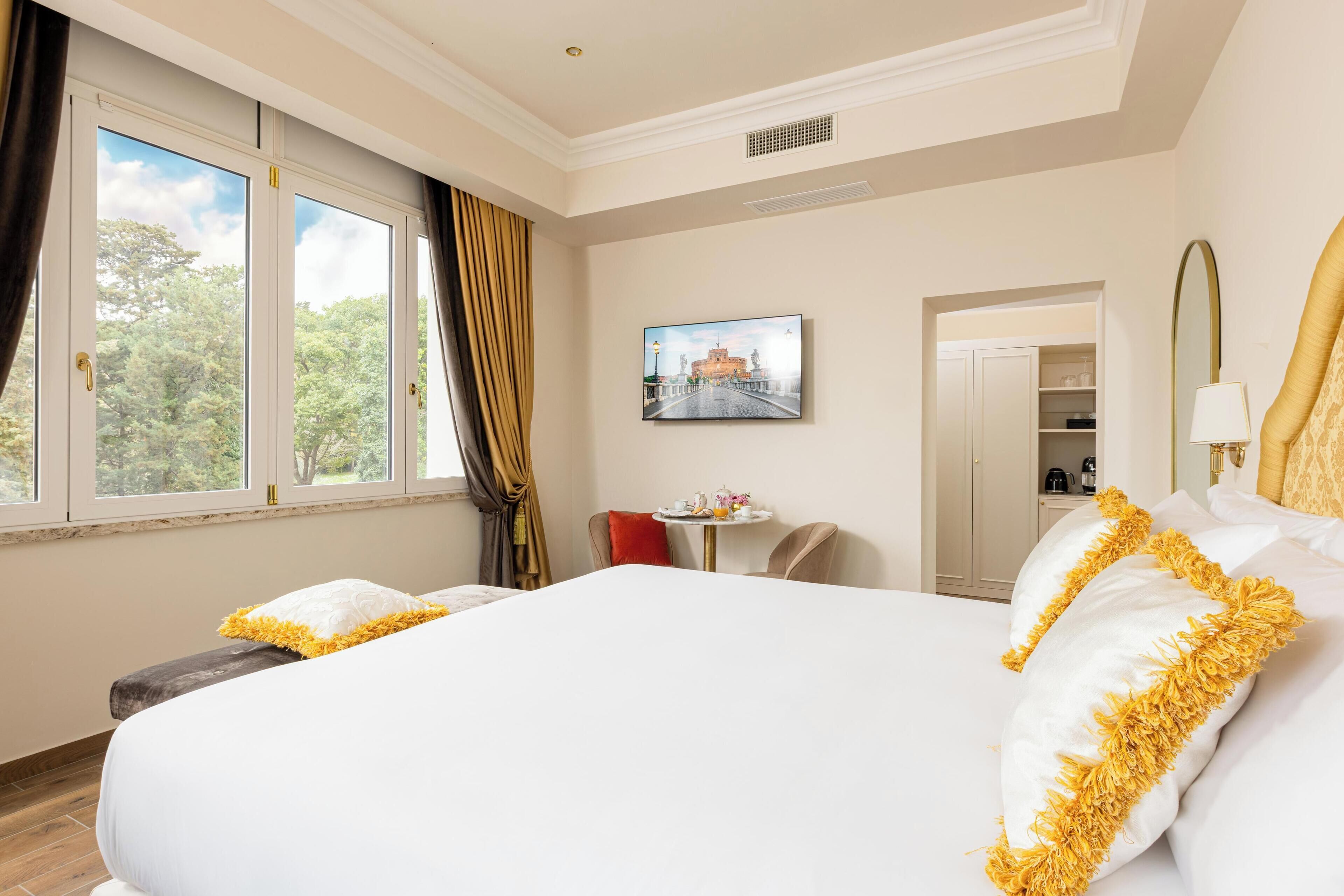 A hotel room with a large bed, a small table with a lamp, a painting on the wall, and a view of trees through the window.