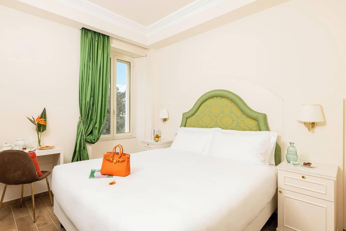 A hotel room with a bed, a desk, a chair, a window with green curtains, and a nightstand with a lamp.