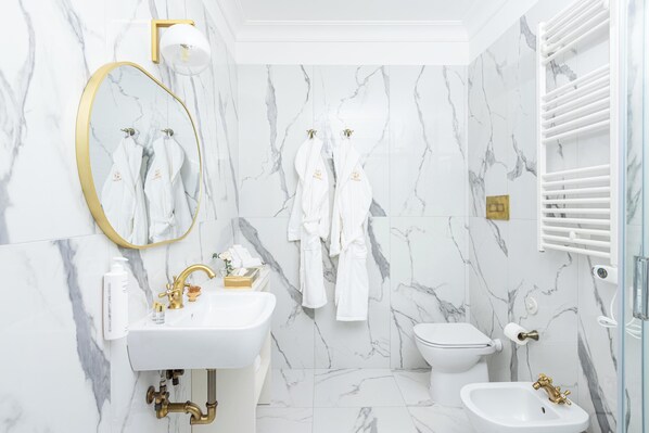 Une salle de bain avec un mur en marbre, un miroir encadré d’or, un lavabo blanc, un robinet doré, des toilettes blanches et un bidet blanc.