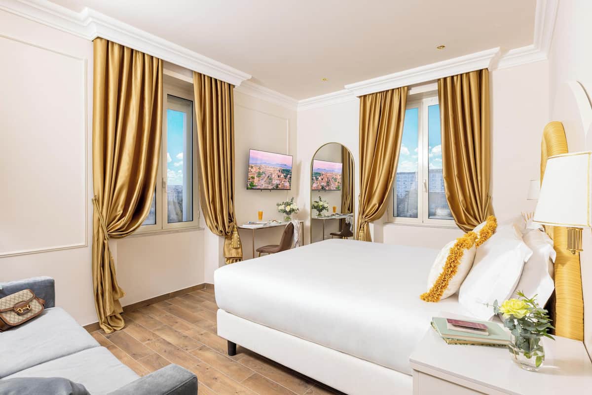 A hotel room with a large bed, a desk, a chair, a sofa, and a TV on the wall.