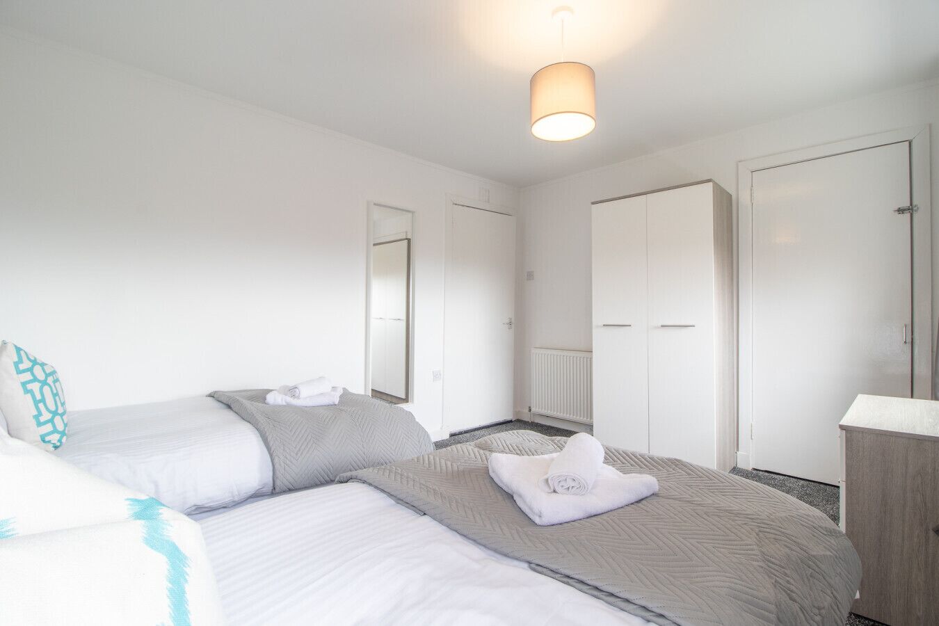 A hotel room with two beds, a wardrobe, a radiator, and a hanging light fixture.