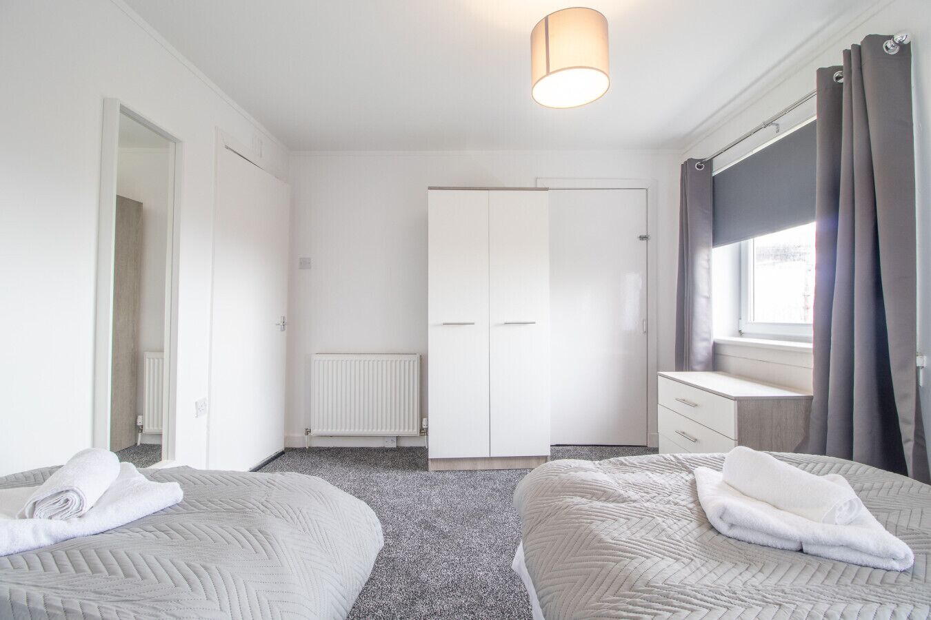 Ein modernes, helles Schlafzimmer mit zwei Betten, einem Kleiderschrank und einem Fenster mit Vorhängen.