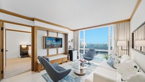 Ein modernes Hotelzimmer mit großem Fenster, einem Flachbildfernseher, einer Couch, zwei Sesseln, einem Couchtisch und einem kleinen Schreibtisch mit Lampe.