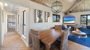 Uma sala ampla com mesa de jantar de madeira, cadeiras de vime, sofá azul e uma televisão grande.